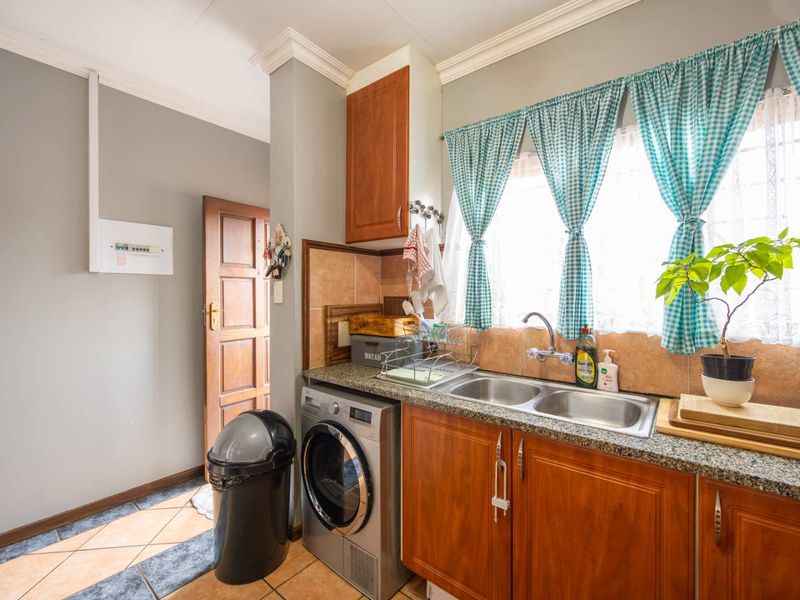 A functional scullery area featuring wooden cabinetry, granite countertops, a double basin sink, and tiled flooring with an integrated laundry appliance space.