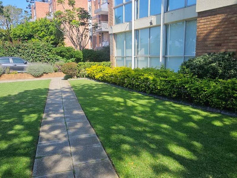 A well-maintained exterior view of an apartment building featuring a brick facade, large windows, a paved walkway, and a manicured lawn.
