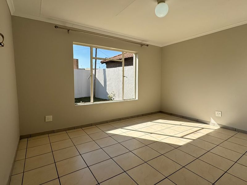 A bright, empty bedroom featuring neutral tiled flooring, a large window allowing natural light, and standard electrical fixtures.