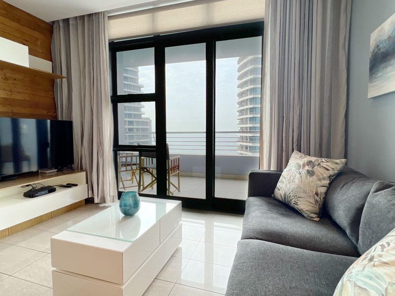 A bright living room featuring large sliding glass doors that open onto a balcony, with modern tiled flooring and a wood-paneled feature wall.
