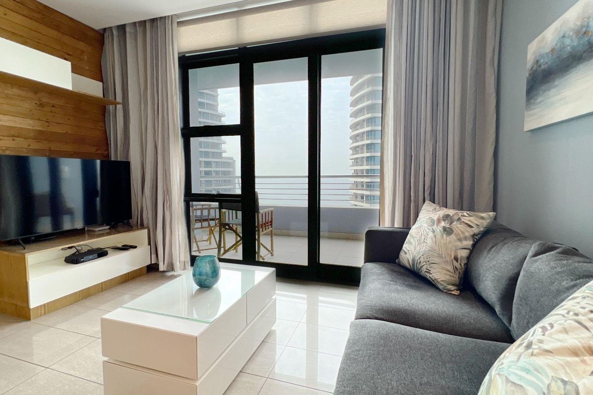 A bright living room featuring large sliding glass doors that open onto a balcony, with modern tiled flooring and a wood-paneled feature wall.