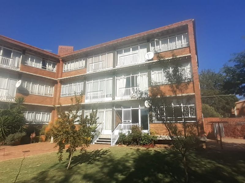 Rear view of a multi-story brick apartment building featuring private balconies, paved courtyard area, and established garden surroundings.