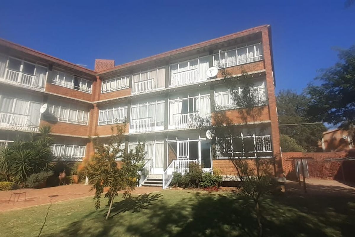 Rear view of a multi-story brick apartment building featuring private balconies, paved courtyard area, and established garden surroundings.