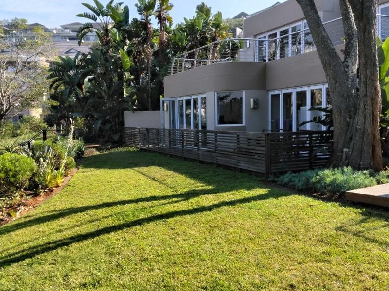 Rear view of the apartment featuring a well-maintained lawn, timber decking, and a modern exterior with large sliding glass doors and stainless steel balustrades.