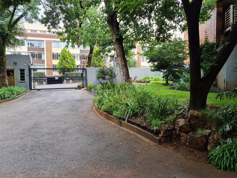 A well-maintained apartment complex entrance featuring an asphalt driveway, secure gated access, and established garden landscaping.