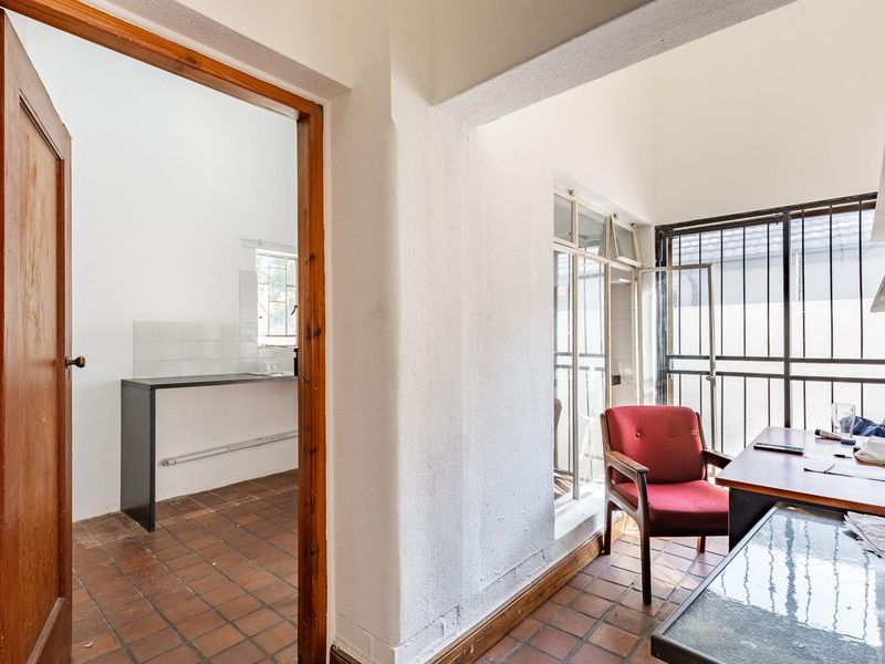 Office space featuring terracotta floor tiles, security bars on windows, and an adjacent kitchenette area with a built-in counter and tiled splashback.