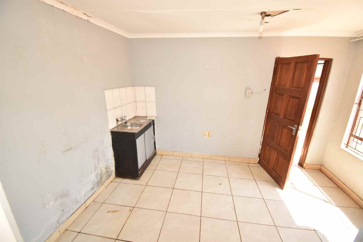 A small, basic kitchenette area featuring a single sink unit, tiled flooring, and a wooden door, currently in need of maintenance and cosmetic repairs.