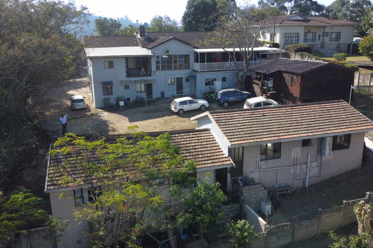 Aerial view of a multi-structure residential property featuring tiled roofs, a large unpaved yard, and established trees in a suburban setting.