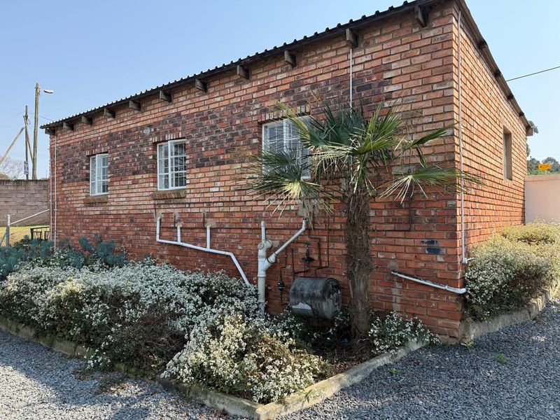Side view of a brick building featuring exposed external plumbing, a geyser, and casement windows set against a landscaped garden bed.