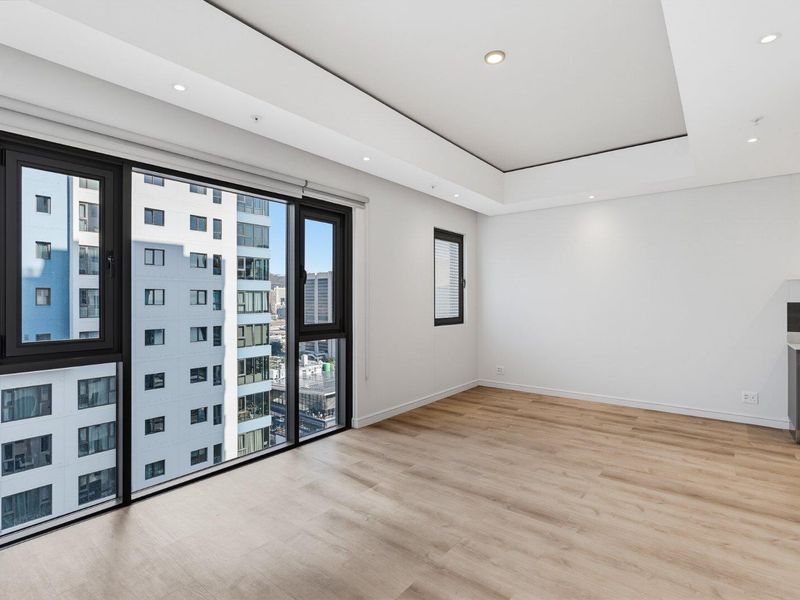 A bright, modern open-plan living area featuring light laminate flooring, recessed ceiling lighting, and large windows with urban views.
