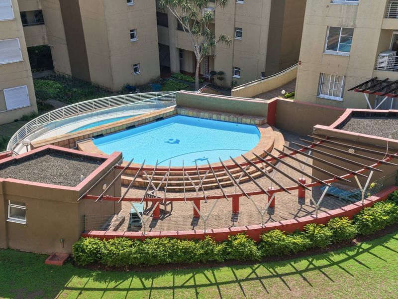 Elevated view of a communal swimming pool area featuring a paved patio, pergola structure, and glass balustrade within a residential townhouse complex.