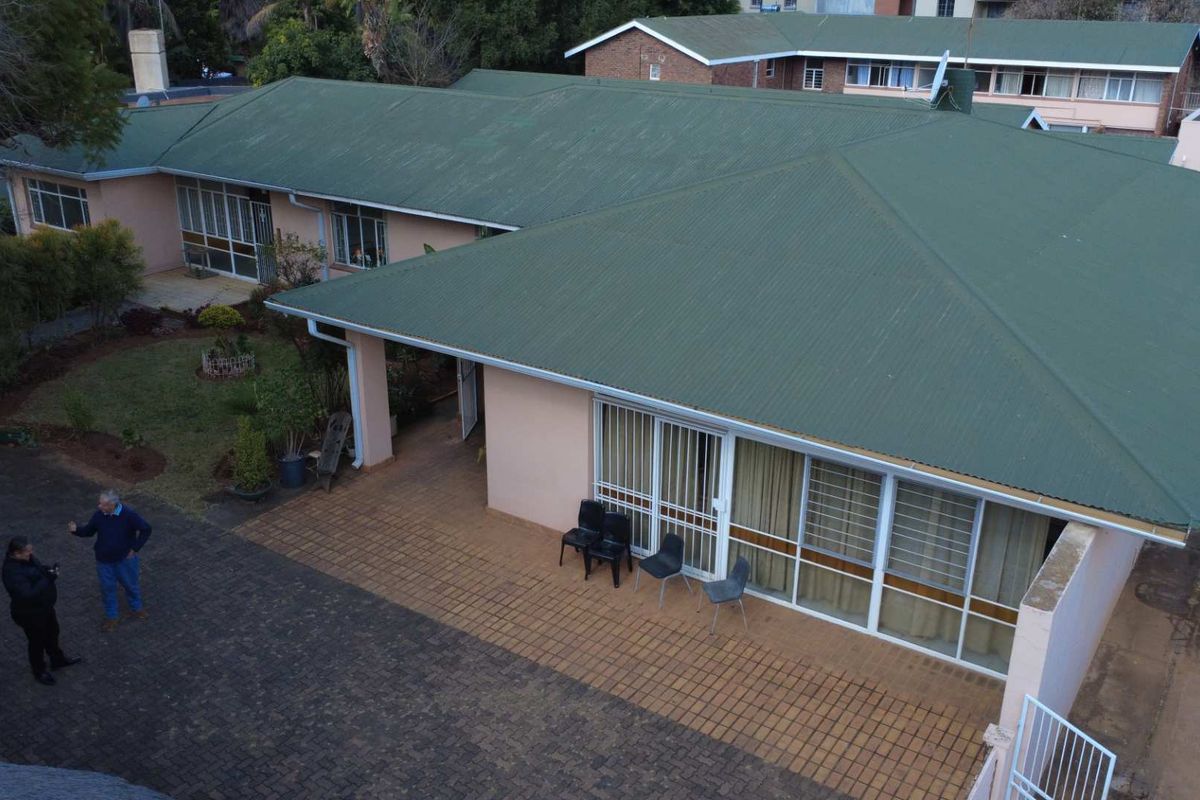 An elevated view of the property featuring a large green corrugated metal roof, paved patio area, and surrounding garden space.
