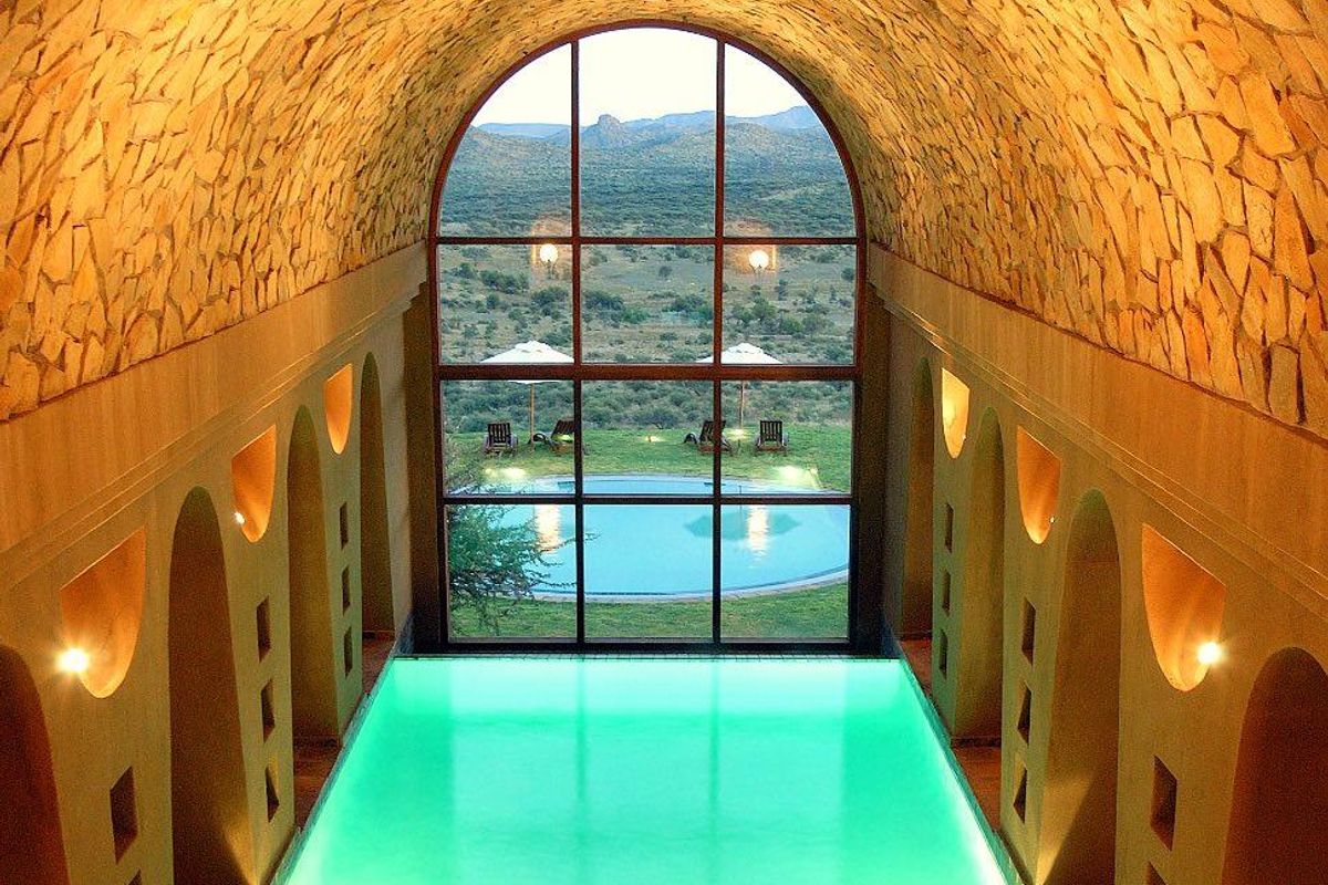 An indoor swimming pool featuring a vaulted stone ceiling, arched wall niches, and a large floor-to-ceiling window overlooking the landscape and outdoor pool area.