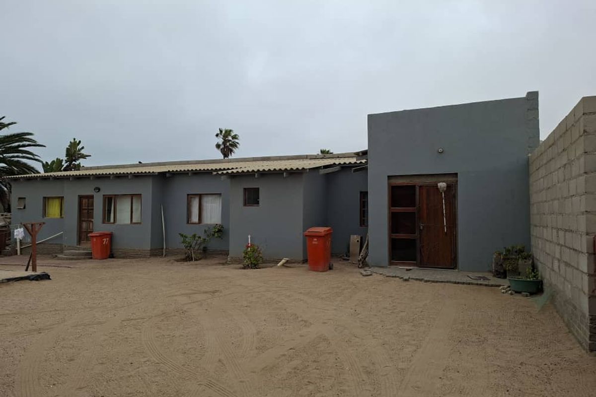 Rear exterior view of a single-story house featuring a grey painted facade, wooden door frames, and an expansive unpaved sandy yard area.