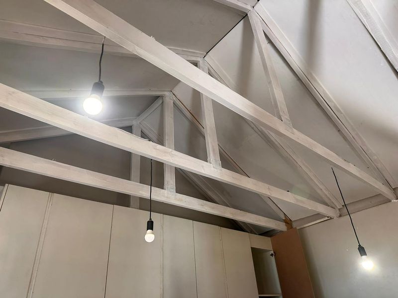 A bedroom interior featuring a high vaulted ceiling with exposed timber trusses, built-in cabinetry, and simple pendant lighting, currently in an unfinished state.
