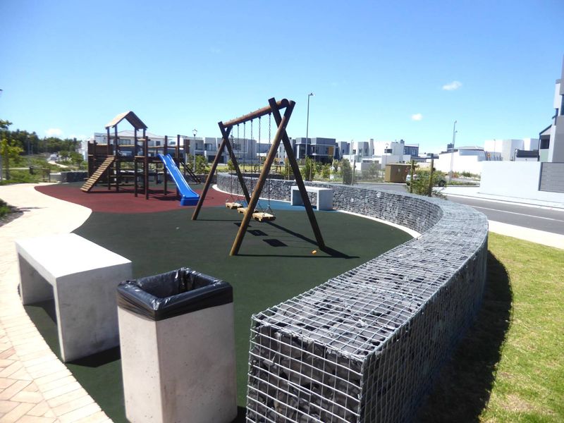 A well-maintained residential playground area featuring modern playground equipment, a curved gabion stone wall, concrete seating, and a durable rubberized play surface.