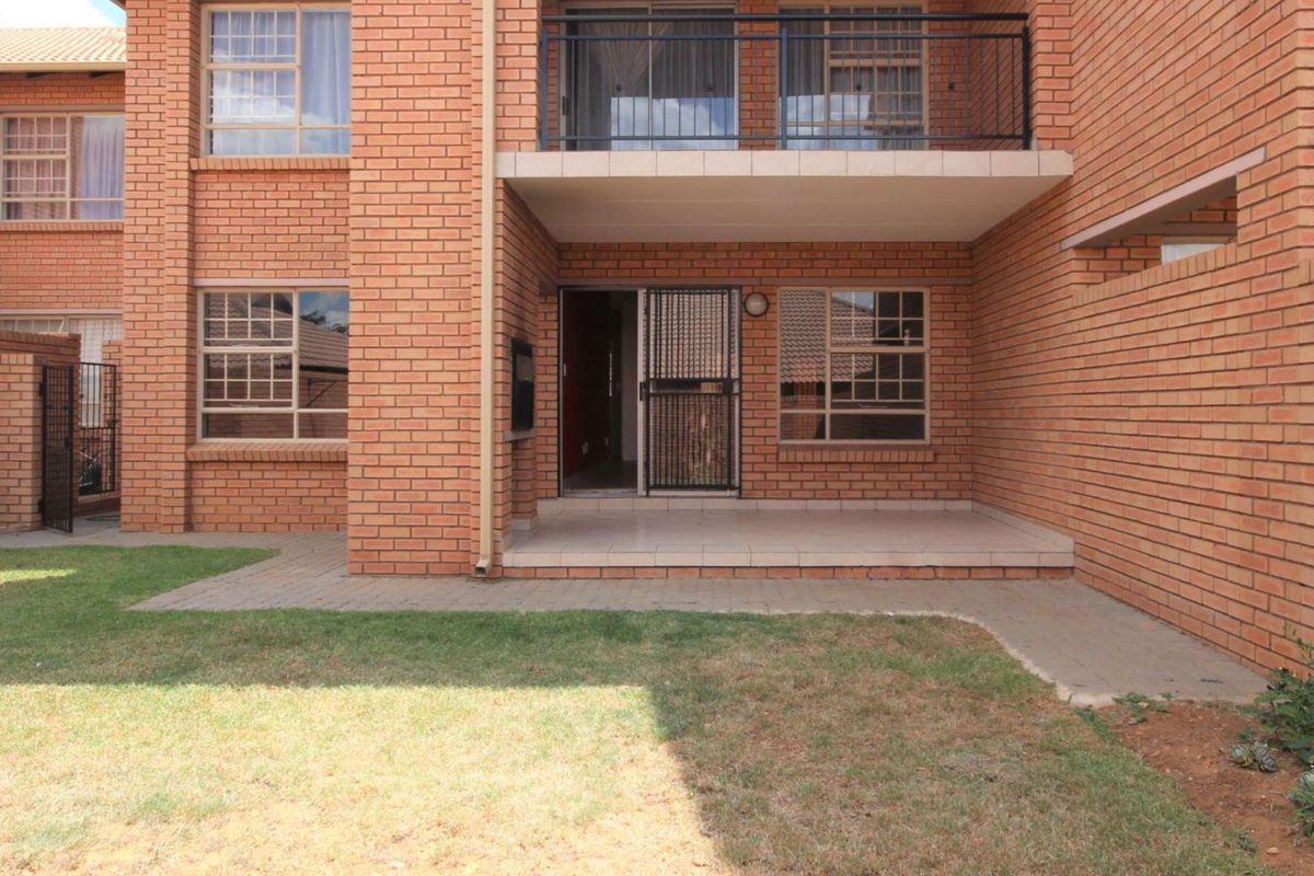 Exterior view of a brick apartment building featuring a ground-floor patio, upper-level balcony with metal railing, and a small private garden area.