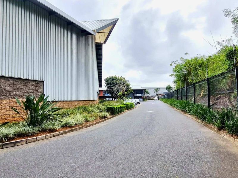 View of the industrial property access road featuring corrugated metal cladding, concrete block masonry, and well-maintained landscaped verges with security fencing.