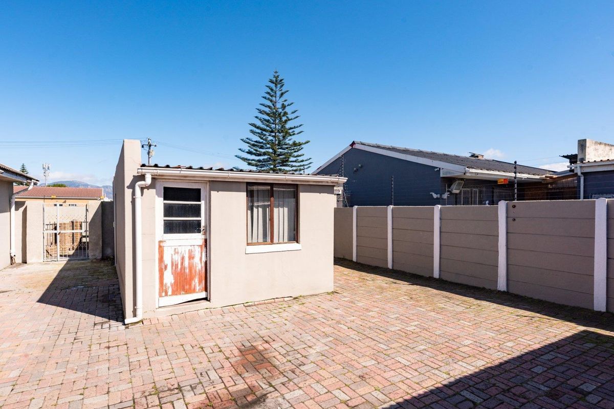 Rear exterior view featuring a paved courtyard, a precast concrete boundary wall, and a small outbuilding with a weathered door.