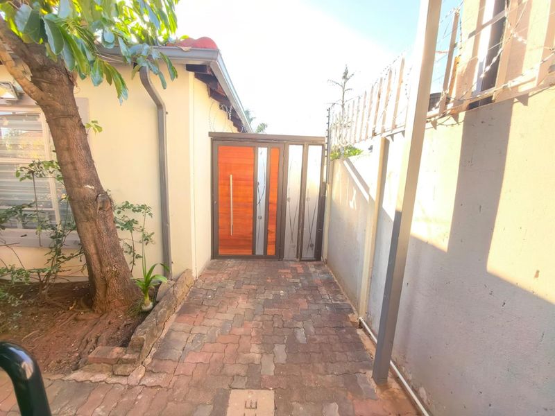 A side entrance featuring a modern wooden door with security gate, paved walkway, and perimeter wall with electric fencing.