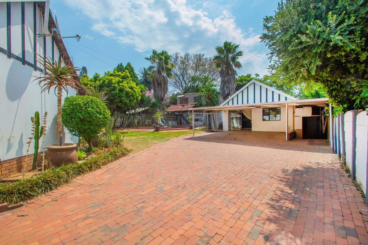 Spacious rear garden area featuring brick-paved driveway, covered patio space, and established landscaping with mature trees.
