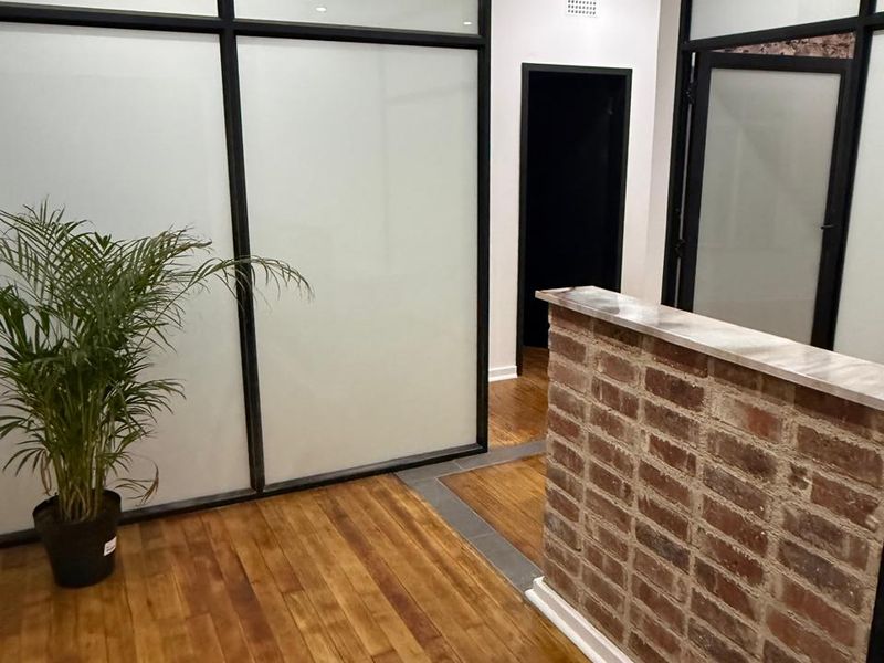 Modern office entry featuring a rustic exposed brick reception counter, hardwood flooring, and contemporary frosted glass partitions with black metal framing.
