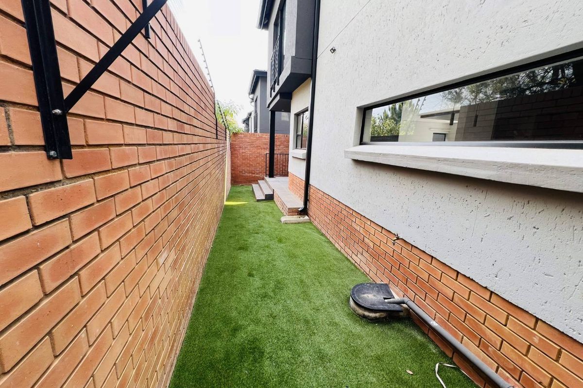 A side passage of the townhouse featuring a brick boundary wall, artificial turf, and modern exterior finishes with concrete steps leading to the rear.