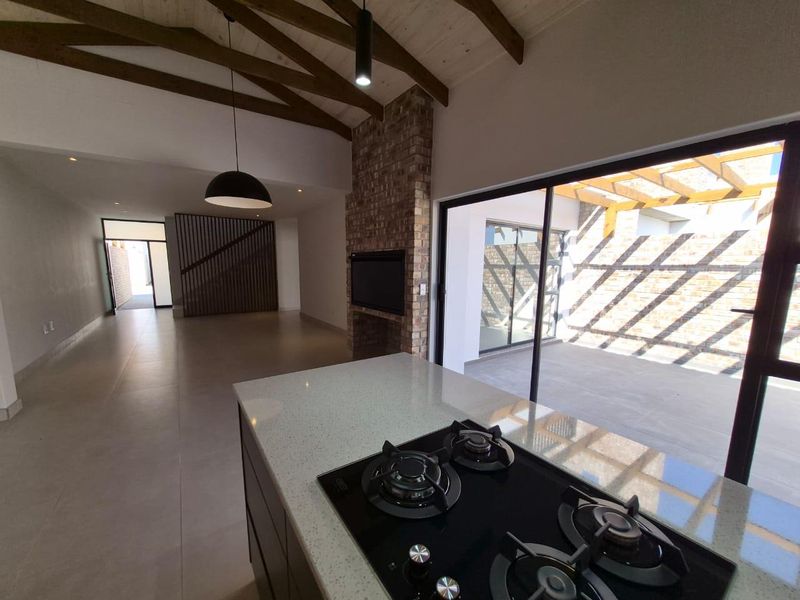 Spacious open-plan living area featuring a modern kitchen island with gas hob, exposed timber beams, and a brick fireplace, opening onto a patio.