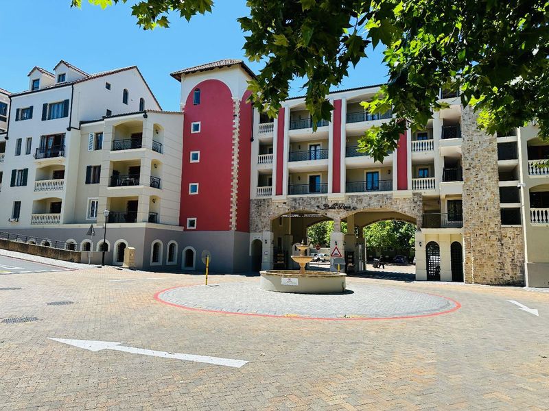 Front exterior view of the Bella Rosa apartment complex featuring a paved circular driveway, central fountain, and distinctive Mediterranean-style architecture with stone and red accents.