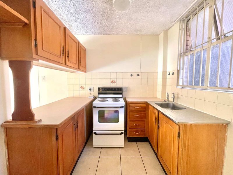 A functional, dated kitchen featuring wooden cabinetry, laminate countertops, a freestanding stove, and a stainless steel sink with tiled backsplash and flooring.