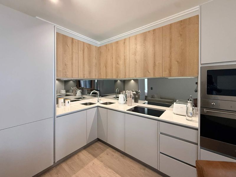 Modern kitchen featuring light grey cabinetry, wood-tone upper cabinets, stone countertops, integrated appliances, and a sleek mirrored splashback.