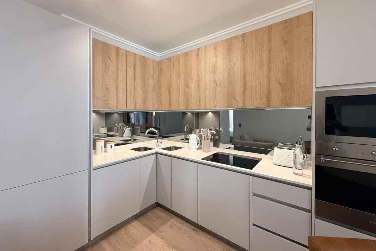 Modern kitchen featuring light grey cabinetry, wood-tone upper cabinets, stone countertops, integrated appliances, and a sleek mirrored splashback.