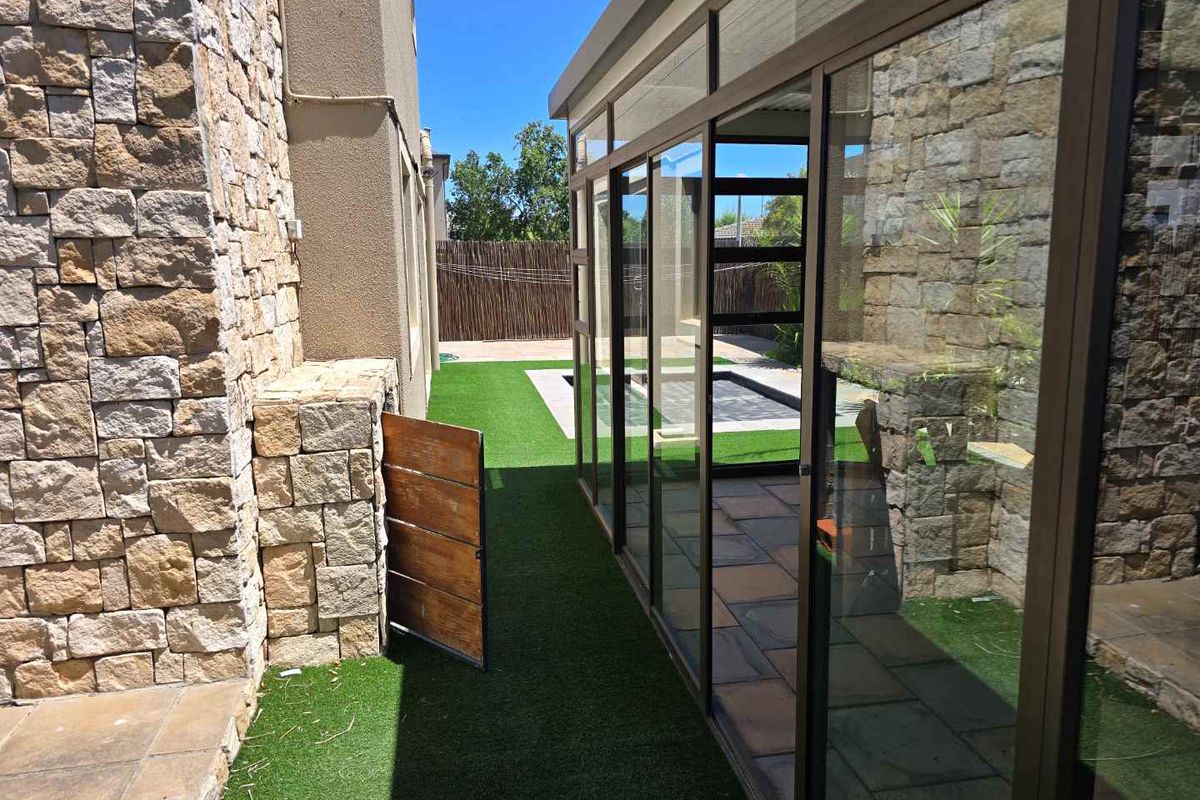 A side exterior view featuring a stone-clad wall, artificial lawn, and a glass-enclosed patio area leading to the rear garden.