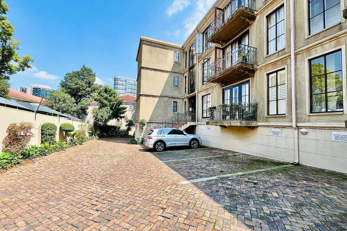Exterior view of an apartment building featuring a brick-paved parking area, multi-level balconies with wrought iron railings, and landscaped garden borders.