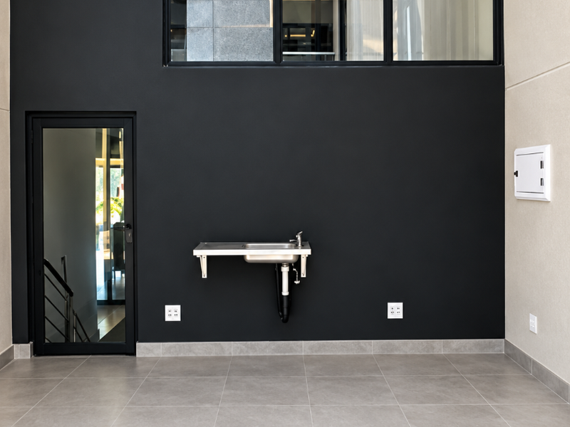 A clean, modern utility space featuring large black-framed windows, tiled flooring, and a functional wall-mounted stainless steel sink.