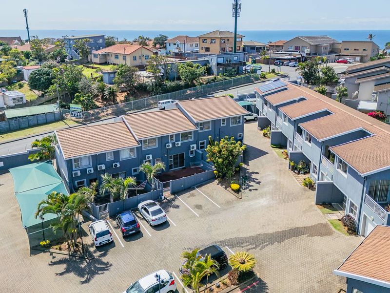 Aerial view of a residential apartment complex featuring paved parking areas, tiled roofs, and solar panel installations within a coastal neighborhood setting.