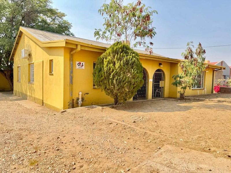Single-story office building exterior featuring a yellow facade, arched entrance with security gates, and a metal roof, set on a gravel lot.