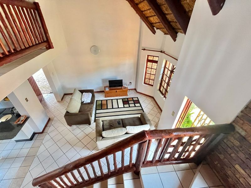 Spacious double-volume living area featuring a traditional thatched roof, exposed timber beams, tiled flooring, and a wooden staircase leading to a mezzanine level.