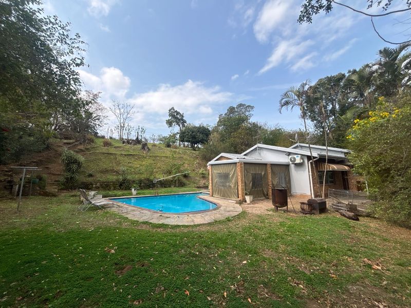 Rear garden view featuring a swimming pool with stone coping, a covered patio area, and a brick dwelling set against a sloped landscape.