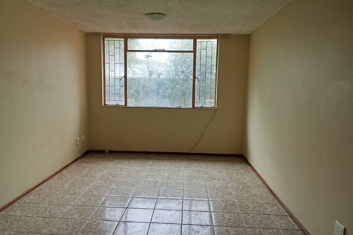 An empty, neutrally painted living room featuring tiled flooring and a large window with security bars, offering a blank canvas for renovation.