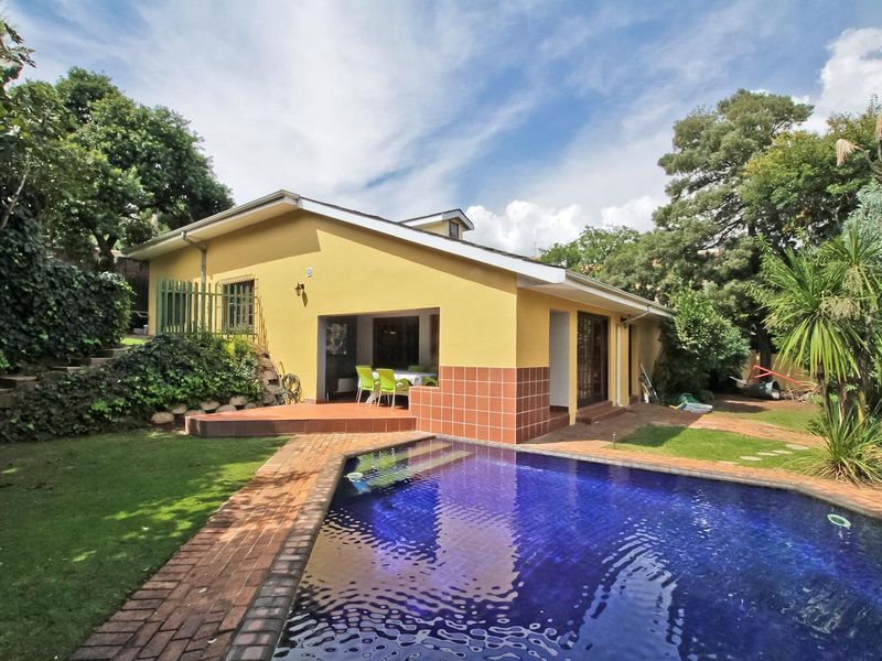 Rear exterior view featuring a swimming pool, brick-paved surround, and a covered patio area set within a landscaped garden.
