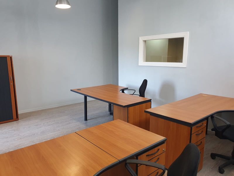 A functional office space featuring light grey walls, laminate flooring, and an internal window providing natural light flow.