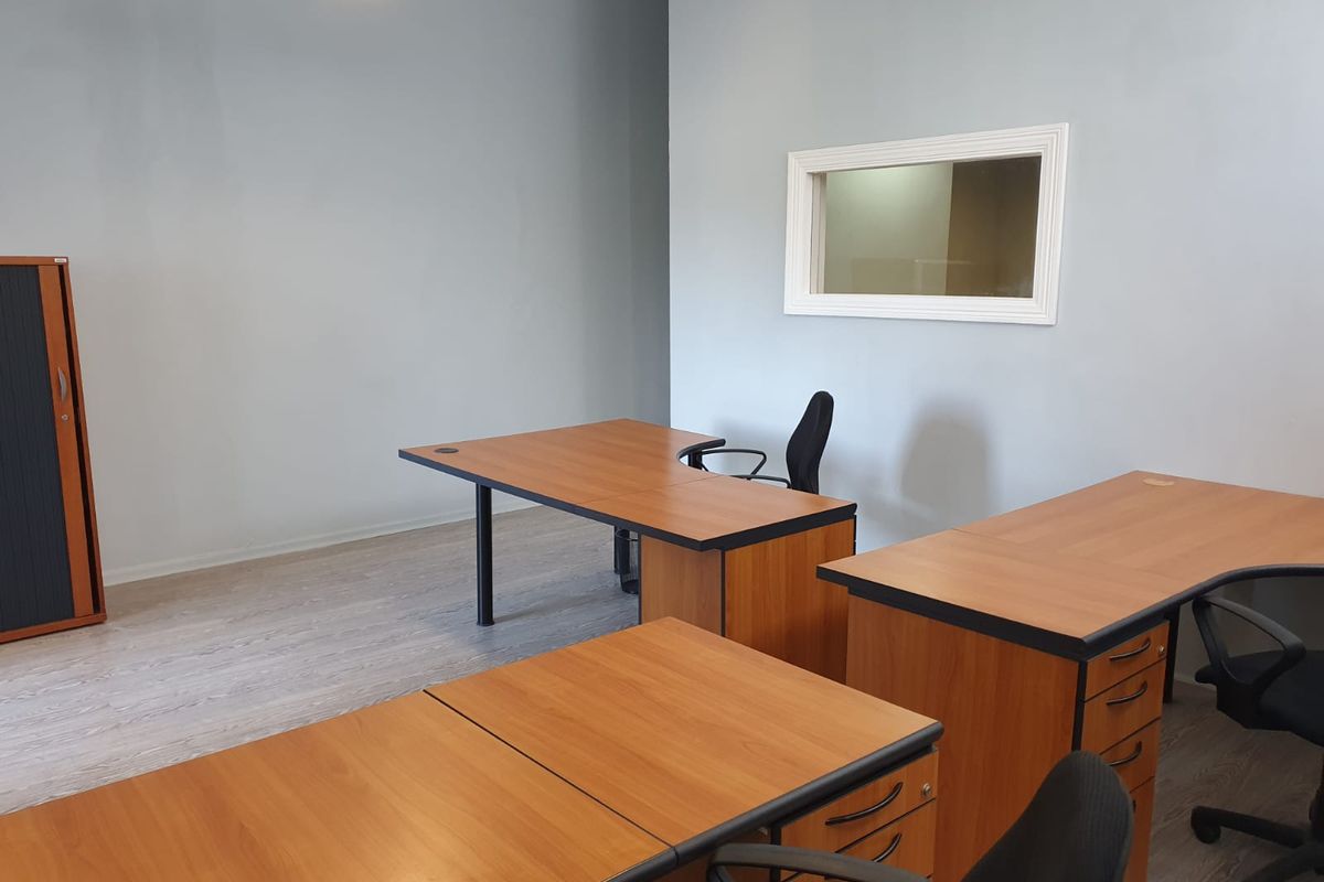 A functional office space featuring light grey walls, laminate flooring, and an internal window providing natural light flow.