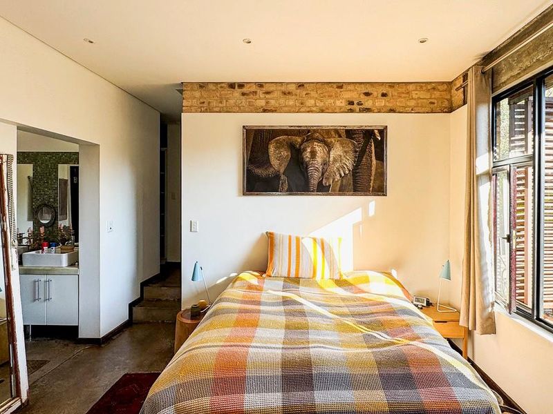 A well-lit bedroom featuring exposed brick detailing, polished concrete floors, and large windows, with direct access to an adjoining en-suite bathroom.
