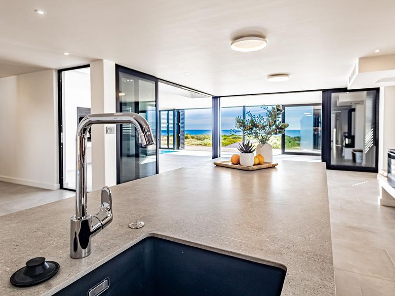 Modern open-plan living area featuring a sleek kitchen island with undermount sink, contemporary fireplace, and expansive sliding doors opening to a coastal view.
