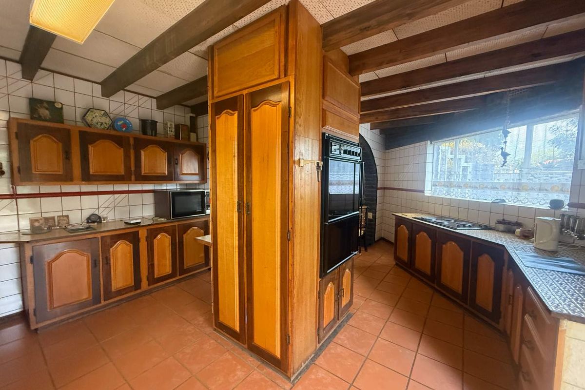 A dated kitchen featuring wooden cabinetry, a central built-in oven unit, tiled countertops, and exposed timber ceiling beams.
