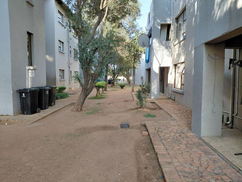 View of the communal outdoor area between apartment blocks, featuring paved walkways, mature trees, and shared refuse storage space.