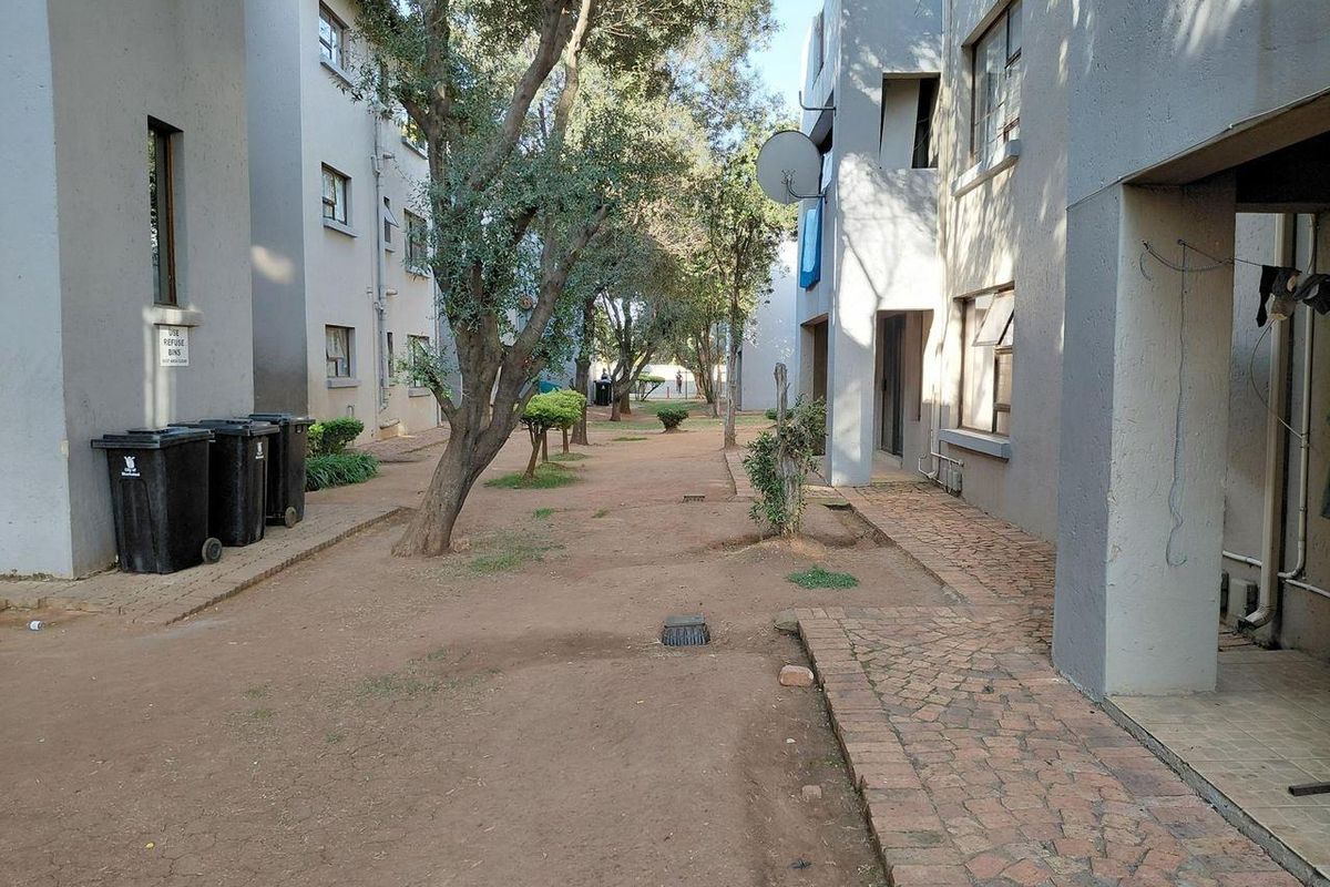 View of the communal outdoor area between apartment blocks, featuring paved walkways, mature trees, and shared refuse storage space.