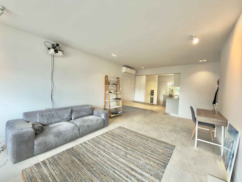 An open-plan living and kitchen area featuring neutral tiled flooring, recessed lighting, and a wall-mounted air conditioning unit, leading into a modern kitchen space.