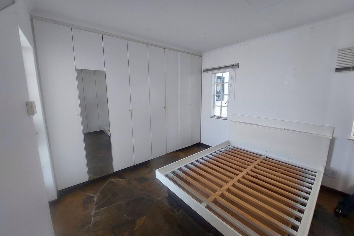 A bedroom featuring built-in white cupboards with a mirrored panel, stone tile flooring, and a small casement window.
